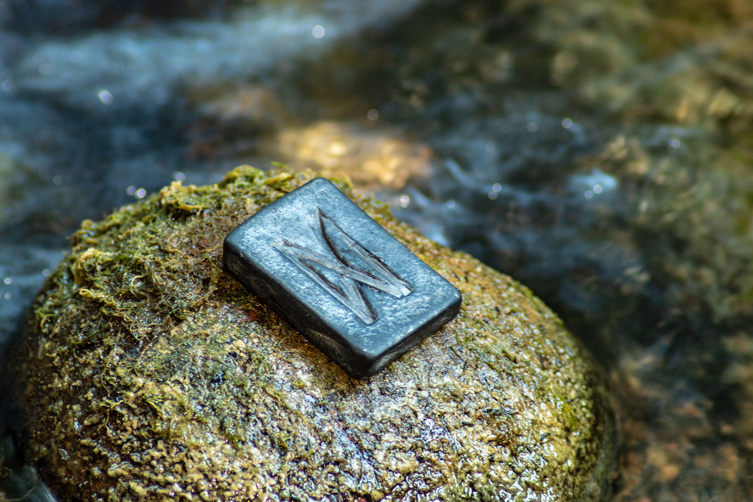 Norse rune Dagaz (Dagr) on a moss covered stone at the edge of a river seen in the background. Dagr is the symbol of Dawn and signifies transformation and quick changes to the good. Some people think it looks like a butterfly.