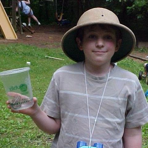 A young boy with a hat on and a frog caught in a plastic container.